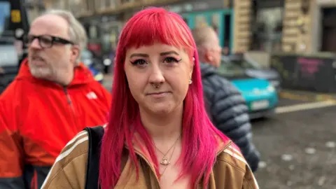 A woman with pink hair stands in the middle of the street with a light brown Adidas coat on