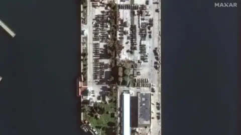 Maxar Military vehicles at the port of Tartous on December 17. They are parked in rows with green buildings surrounding them.