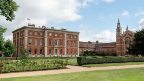 A large square three-storey red-bricked historic school building is in the foreground, with a larger building with spires and a courtyard in the background. A green field and a topiary hedge provide a border.