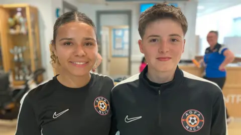 Two women wearing Nike-sponsored black football attire. One has tied-back brown hair and the other has short brown hair.