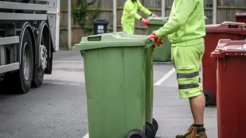 A council worker in green high-vis clothing and red gloves holds on to the handles of two green waste bins on a street with a bin lorry nearby.