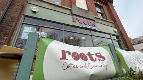 The exterior of a coffee shop called Roots. It is a brick, end of terrace building with olive green windows and a sign with pink writing on saying "Roots". There is a red, green and beige banner outside the café, bearing its name. The sky above it is wintry and cloudy.