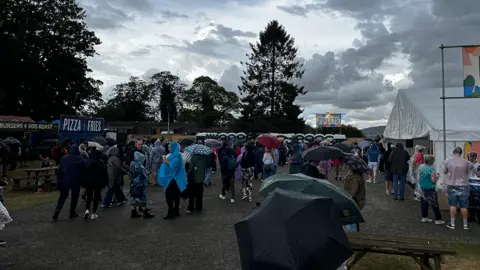 Crowds of people at a festival. Many are holding umbrellas or wearing raincoats. Food and drink stalls are on the left and right, with portaloos in the background