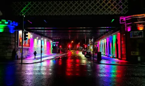 Ruth Cumming A city street on a wet and dark night. Various lights - purple, red, green and blue - are reflecting across both sides of the road. 