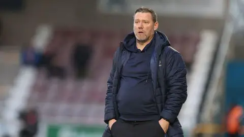 Getty Images A man in a navy sports coat and navy hoodie stands with his hands in his pockets in a football stadium.