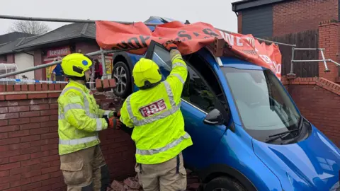 Two firefighters are pictured with a car hanging over a wall after a crash in a supermarket car park