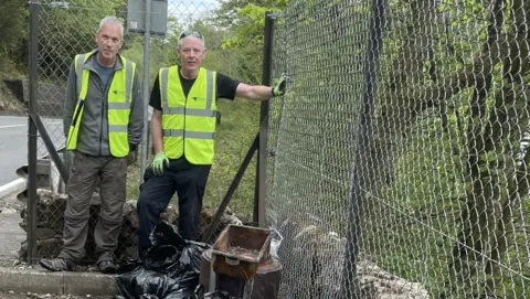 Two men stand behind a bunch of abandoned bags, a broken metal gate and some rusty old metal that have been abandoned in a layby