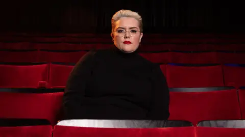 Comedian Jayde Adams sitting in an empty theatre with a solemn look on her face, wearing a black turtle neck jumper and slicked back hair.