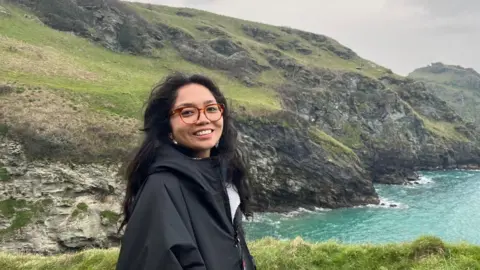 Supplied A young woman wearing glasses and a black jacket smiles while standing on a grassy hill overlooking the ocean and rocky cliffs. 