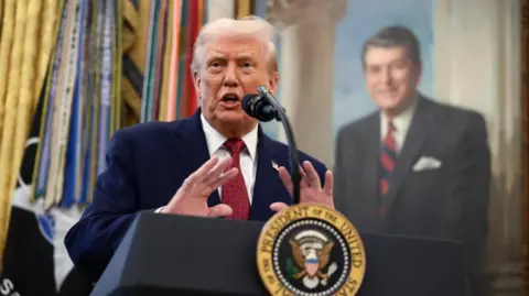 Reuters U.S. President Donald Trump speaks at a lectern in a navy suit with a red tie in front of a painting and flags