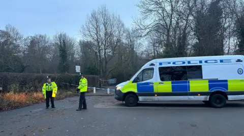 BBC A blue and white police cordon is stretching across a path. There is a white, blue and yellow police van parked in front of it. Two police officers are standing near the van in yellow police jackets and black police hats.