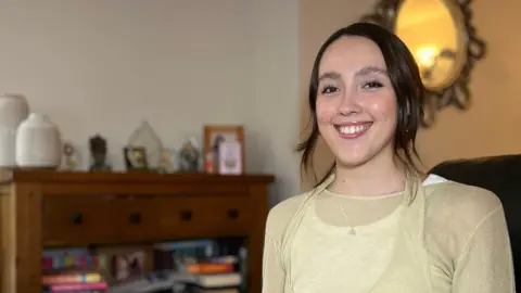 Julia is seated on a dark-colored sofa in a cozy living room. She is wearing a light beige long-sleeve top layered over a lighter shirt and blue jeans, with hands clasped together. Behind Julia, there is a wooden sideboard with multiple drawers, decorated with framed photos, ornaments, and books. To the left, a tall gold planter holds a white orchid, and a large decorative mirror hangs on the wall above the sideboard. The room has warm lighting and a neutral color palette.