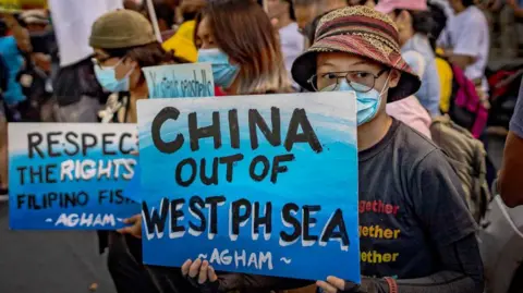 A proester in Manila, wearing a hat, eyeglasses and face mask carrying a blue sign with the words China out of West Philippine Sea in bold black font