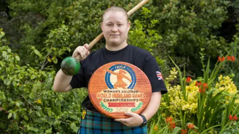 BarronMedia Eve is wearing a green and blue kilt and holding a wooden disc with Woman's Amateur World Highland Games Championships 2025 engraved on it.