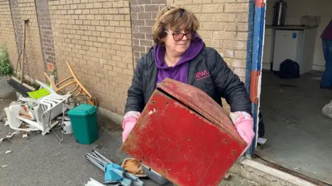 A woman with a brown, medium bob, wears glasses, a black coat and purple sweatshirt with pink marigold gloves on and holds a red box outside the clubhouse. 