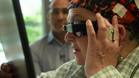 Sheila is wearing the special glasses, reading from a tablet a few centimetres from her face - she is side-on, holding one hand to the side of her face and concentrating hard. Watching behind her, slightly blurred, is consultant ophthalmic surgeon Mahi Muqit, from Moorfields Eye Hospital.