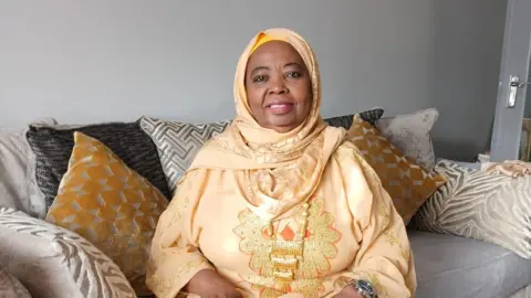 A woman sitting on a sofa wearing a headscarf and an embroidered gown.