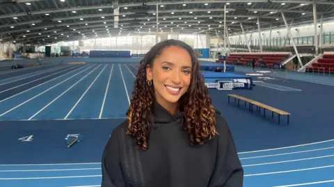 Laviai is sat down at an indoor track. She is wearing a black hoodie, she has dark brown curly hair and gold hoop earrings. The track behind her is blue with white lines. 