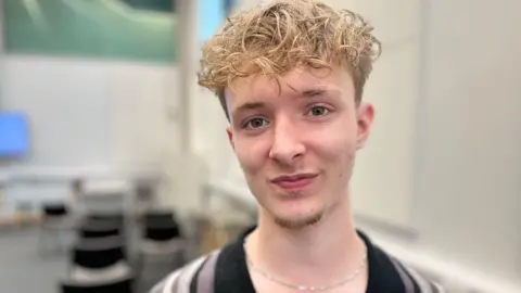 A young man with curly blond hair and a silver necklace chain looks directly at the camera. He is standing in a lecture room with chairs and a screen behind him. 