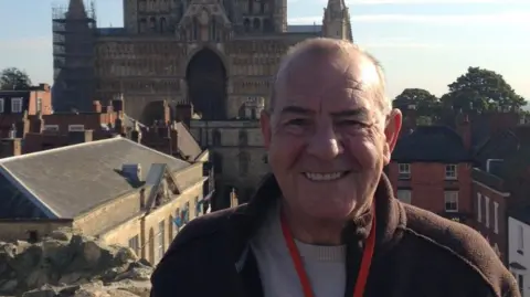 Jenny Fox A head and shoulder photo of Jenny's husband, Alan, stood smiling at the camera. He has very short, balding hair and is wearing a light grey sweater, a brown fleece and a red lanyard. Behind him in the distance is Lincoln Cathedral. 