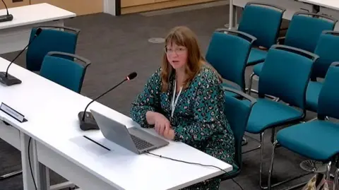 Cambridgeshire County Council Screengrab of a YouTube livestream from Cambridgshire County Council. Bryony Rothwell is sat at a white table with a laptop and microphone in front of her, and some turquoise chairs behind her. She has brunette hair, is wearing glasses and a turquoise dress.