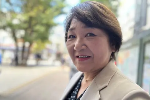 A Japanese woman wearing a blazer and a blouse looks at the camera