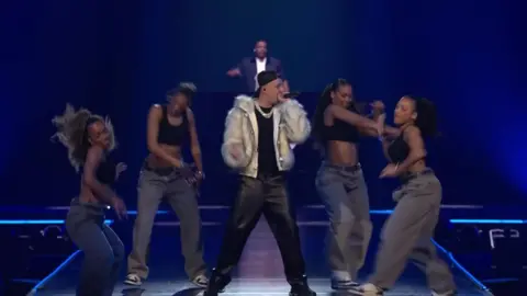 Aitch, wearing a white fake fur jacket and black leather trousers, sings on stage at the MOBOs surrounded by four dancers wearing grey trousers and black crop tops. The stage is lined with blue strip lights.