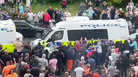 A riotous crowd gathered around a police van, with several members of the group pushing up against the van making it wobble, taken from inside the Rotherham Holiday Inn Express on 4 August