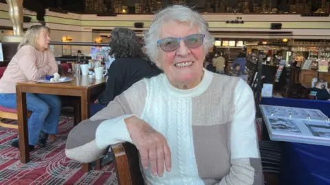 A woman in her late 80s sits turned sideways in her chair at a pub table, with her arm wresting on the back of it. She is wearing a white and beige jumper with a block pattern on it and blue-rimmed glasses with tinted lenses. She has short grey hair.