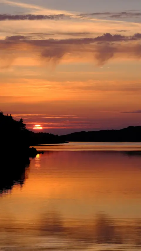Julian Gillespie A tranquil sunset over a calm body of water. The sky is a vibrant mix of oranges, pinks, and purples, with soft, flowing clouds. 