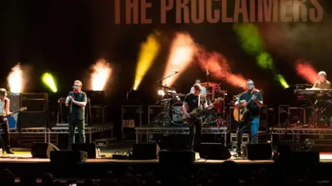 Getty Images The Proclaimers are playing on stage, with the brothers singing into mics with two guitarists alongside them, with a keyboardist and a drummer on separate stage risers. There are various coloured stage lights, projecting through smoke and a sign The Proclaimers above them. 