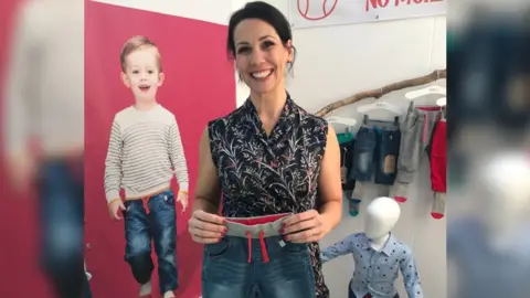 Natalie Hull Natalie Hull standing in front of a Sockatoos advert in a black top and holding a pair of the sockatoos trousers