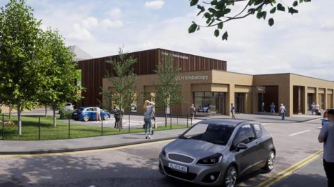 A computer-generated image of the proposed endoscopy unit at Lincoln County Hospital. The building is split into a single-story, light brown section and a two-story dark brown section. It is fronted by a small car park and road, with a car passing, and landscaped grounds, including a leafy green tree.