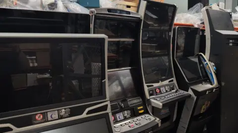 A row of electronic gambling machines stored closely together, most with large black screens and control panels featuring rows of buttons. In the background are shelves and stacked items, including cardboard boxes and plastic-wrapped materials.