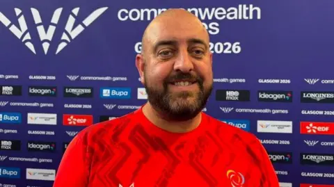 Manx Radio Lee Holland, wearing a red Isle of Man team top, stands in front of a media booth board, on which it says 'Commonwealth Games 2026' and has the logos of various advertisers below.