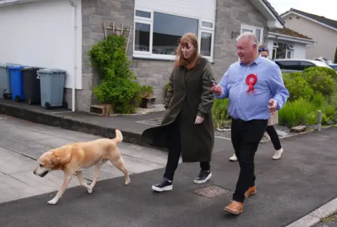 PA Media Davy Russell in a blue shirt alongside a woman with red hair in a long coat with a Labrador dog walking on a residential street