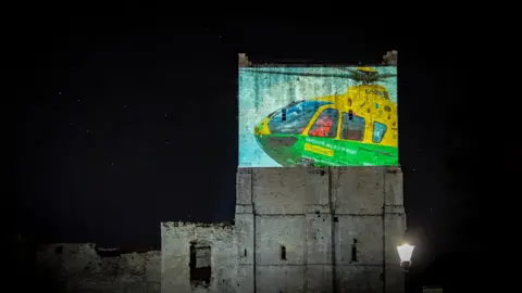 HIOWAA An old castle building at night time. It is tall and in the top half, an image of a green and yellow helicopter is projected on to it.