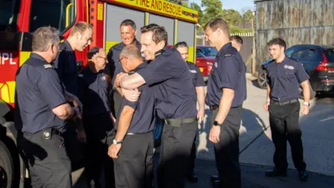 HIWFRS The Minall twins being hugged by other firefighters at the station near a fire truck. Everyone is smiling. A black car can also be seen behind.