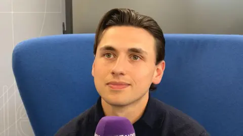 Ryan Maher sitting in a blue chair during an interview. He is holding a microphone and looking away from the camera.