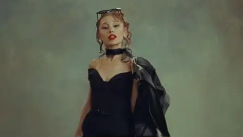 Babydoll Music/Republic Records/YouTube Ariana Grande in a black dress with a black biker jacket over one shoulder and dark glasses on her head.