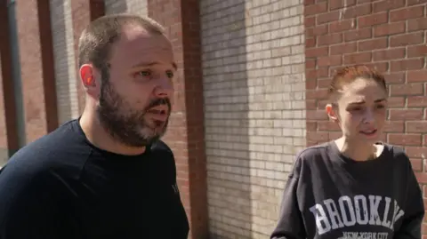A man with a shirt beard and a woman with red hair stand speaking in front of a brick building.