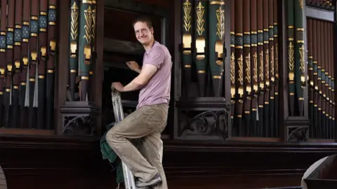 A man in cream jeans and a pink T shirt is pictured climbing a ladder and smiling at the camera before climbing inside an historic pipe organ to start restoration work. 