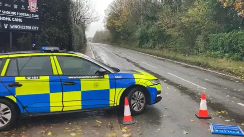 A police car is parked at the side of a road near some traffic cones. A 'police incident' sign has fallen over.