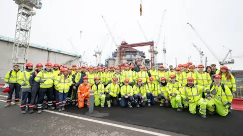 Aran Jefferies A large group of people wearing high-vis gear and hard hats pose outside of Hinkley Point C. More than a dozen cranes can be seen in the background as well as the top of the nuclear reactor, which is a dome structure.