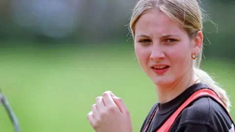 Getty Images A headshot of a blonde woman looking to the right of the camera. She has straight hair tied back in a low ponytail and gold earrings and necklace. The grass of a football pitch can be seen behind her.