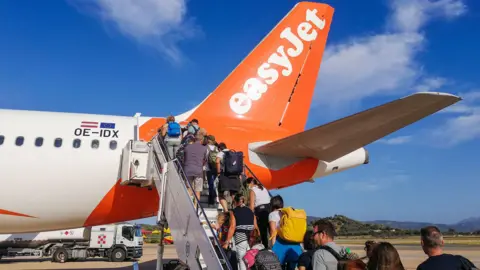 People boarding an EasyJet flight some carrying hand luggage and rucksacks on airport tarmac in Sardinia. It's a sunny day and the sky is blue.