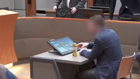 Reuters A man in a jacket sits in a courtroom looking at an image of a mountain, wearing a blue jacket