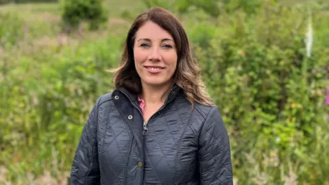 A woman with shoulder-length brown hair smiles at the camera with green plans behind her. She is wearing a blue padded jacket.