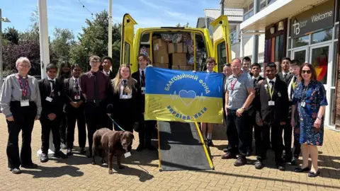 Martin Heath/BBC A line of school pupils along with a female vicar to the left and the school chaplain to the right. Also in the picture is Steve Challen, aid volunteer. They are standing either side of the open doors of a yellow ambulance, which can be seen to be full of medical supplies. There is a blue and yellow flag in front of the ambulance with Cyrillic writing. There is a school building to the right with "Welcome" above the door.