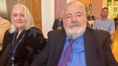 Professor Suzanne Martin and Ivan Martin. Suzanne has blonde shoulder-length hair, wearing a black jacket, black top and lanyard. Ivan has white hair and beard, wearing a black blazer, blue shirt and pink and blue check tie. Other people are sitting in the background.